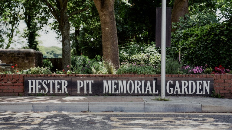 A low wall on the side of a road has flowers and trees behind it. On the wall there is a sign marked Hester Pit Memorial Garden